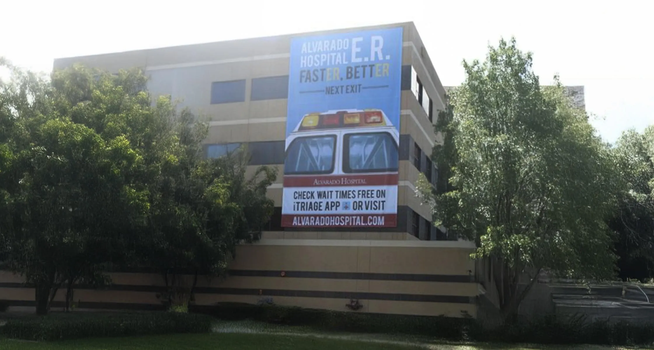 The Alvarado Hospital emergency room banner installed on the side of the hospital building, reading Alvarado Hospital E.R. Faster. Better. Next Exit, with an ambulance graphic, above trees and the hospital entrance