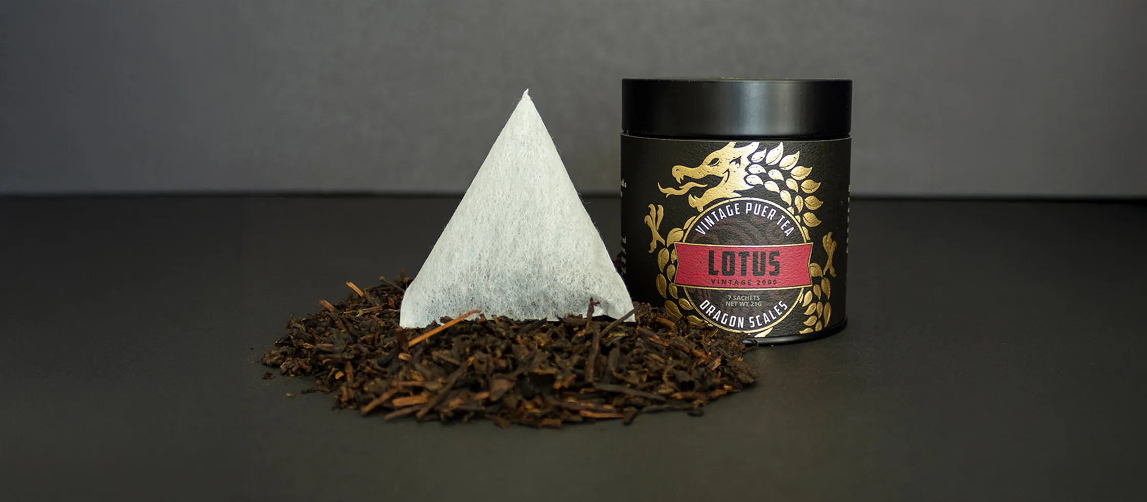 A Dragon Scales pyramid sachet standing in loose Puer leaf beside a Lotus tin