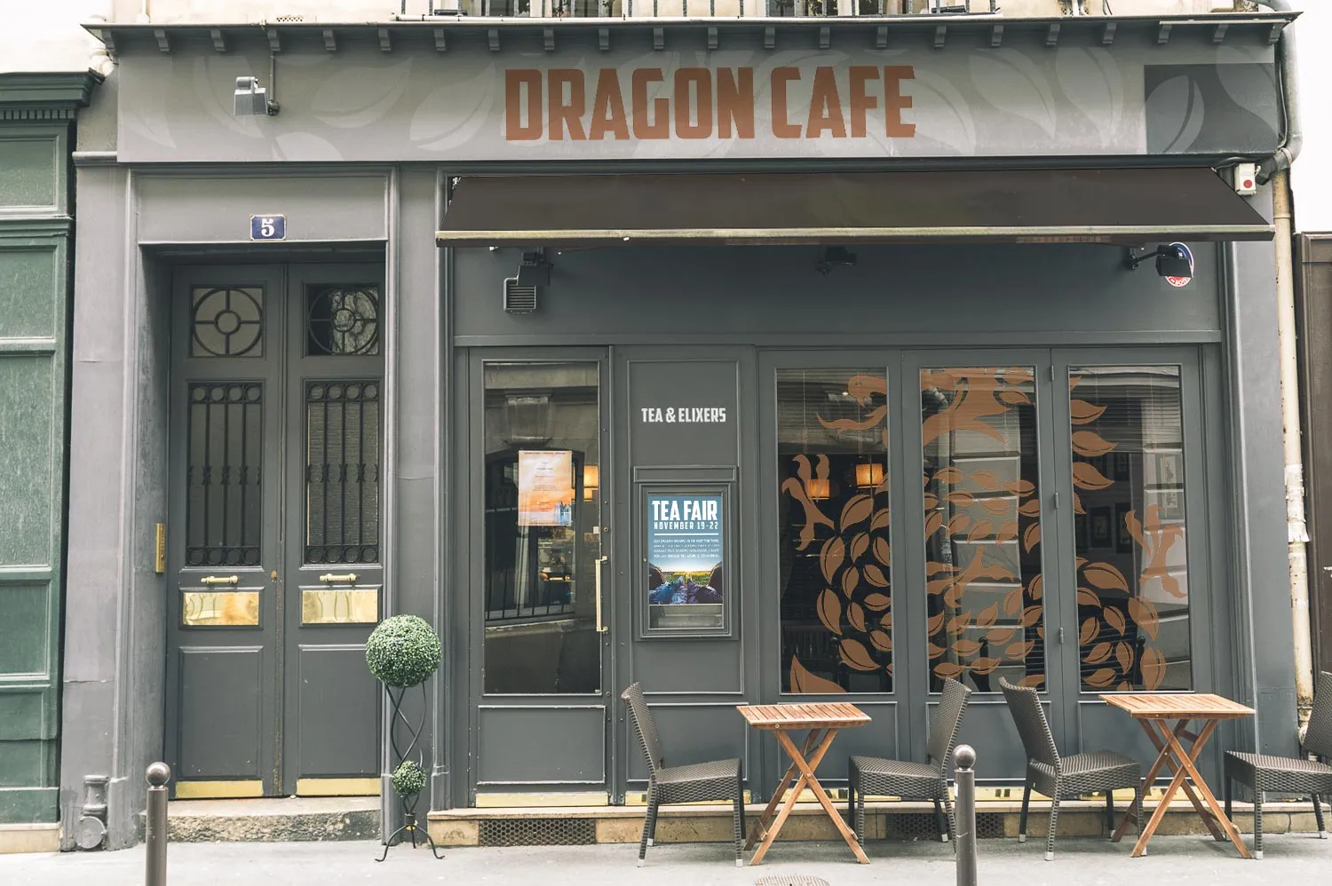 The physical Dragon Cafe tea house storefront, with Dragon Cafe signage, a Tea and Elixers doorway, dragon-scale window graphics, and sidewalk cafe seating