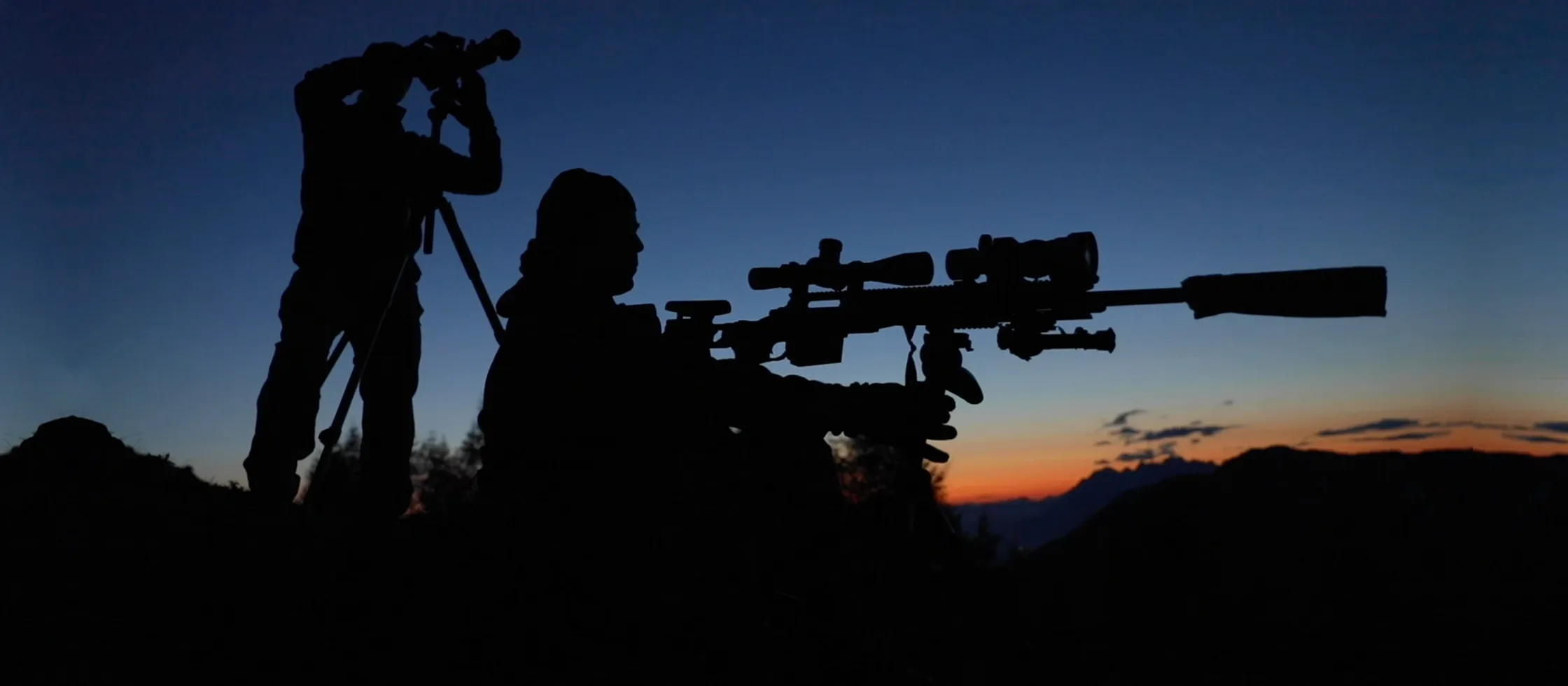 Two figures in silhouette at dusk in a Greystone training environment, a spotter and a long-range rifle against a sunset
