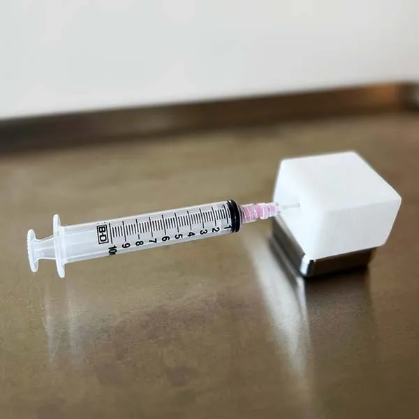 A hypodermic syringe with its needle pushed into the HypoFoam block, the assembled unit resting on a steel surface