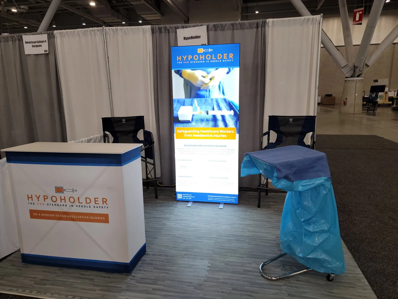 The HypoHolder booth at the American College of Surgeons meeting: the branded counter, a retractable banner headed Safeguarding Healthcare Workers from Needlestick Injuries, and a draped surgical stand