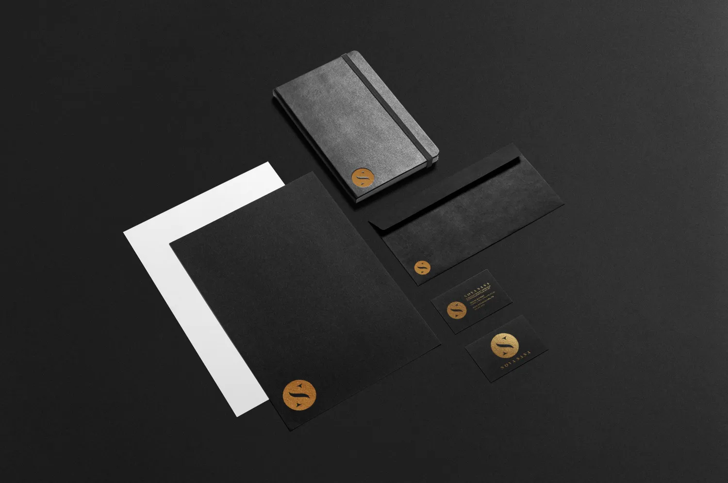 The stationery system on black: a notebook, envelope, letterhead and business cards, each carrying the mark in gold