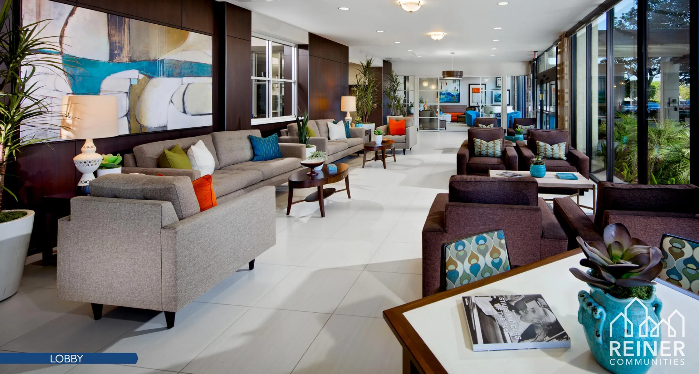 A bright, contemporary Reiner Communities property lobby with modern furniture, artwork, plants, and floor-to-ceiling windows, the Reiner Communities logo in the corner