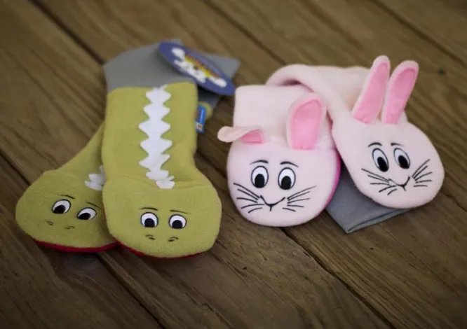 Two pairs of Sputtens sock-puppet mittens on a wood surface: green dinosaur faces and pink bunny faces, one pair still carrying its hangtag